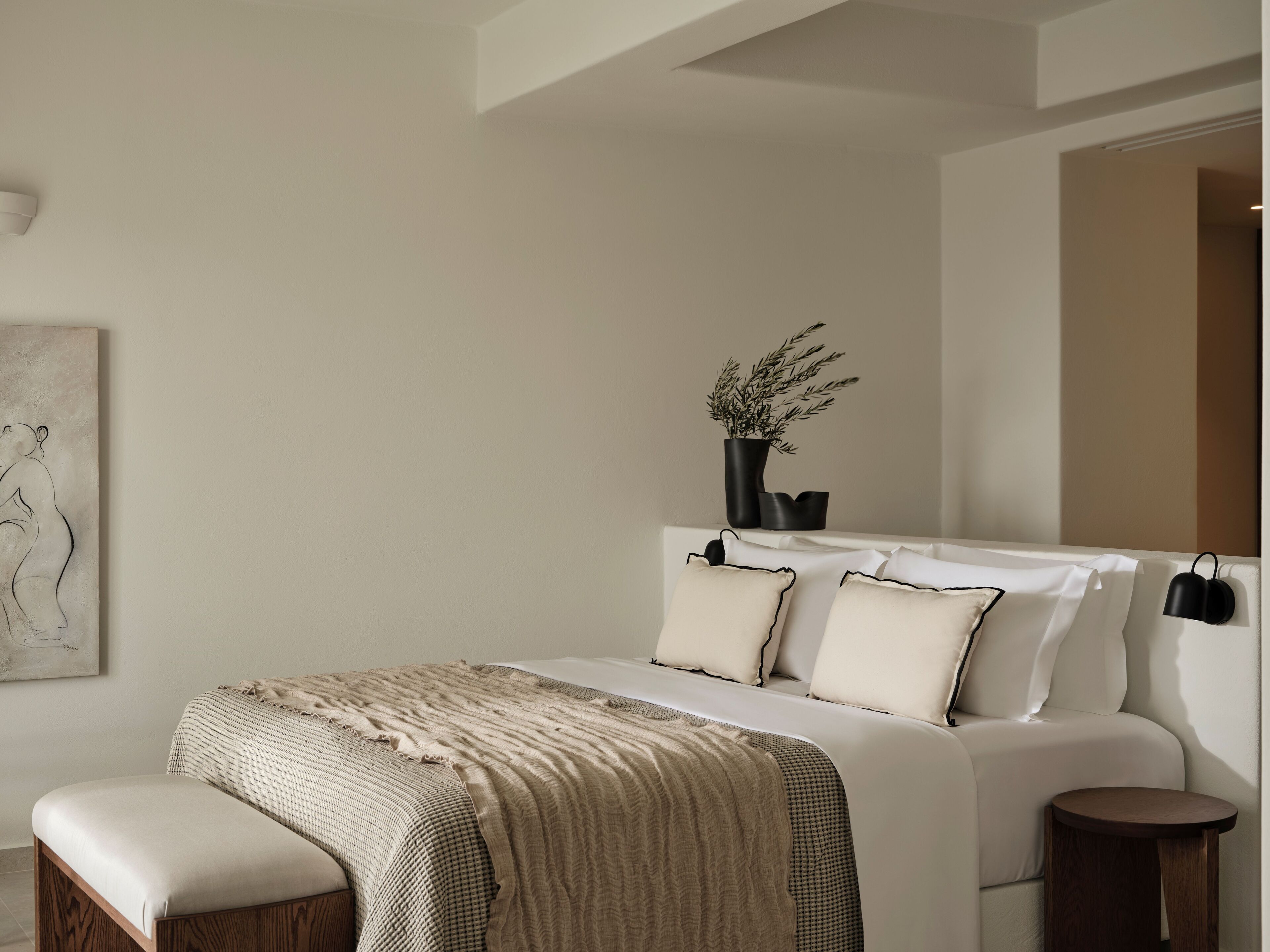 A modern bedroom with a large bed, a bedside table, a small stool, and a wall-mounted lamp.