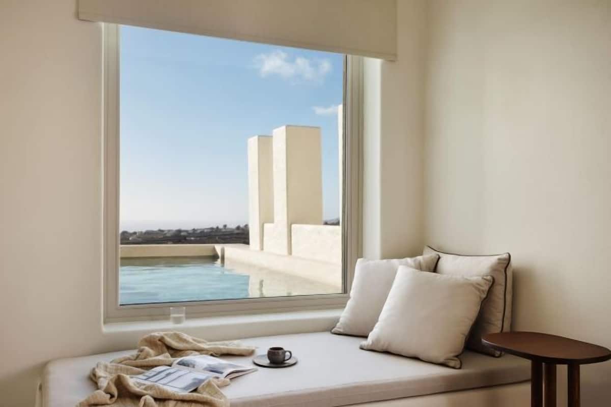 Quarto com janela vista para piscina e edifícios, um sofá com almofadas e uma pequena mesa.