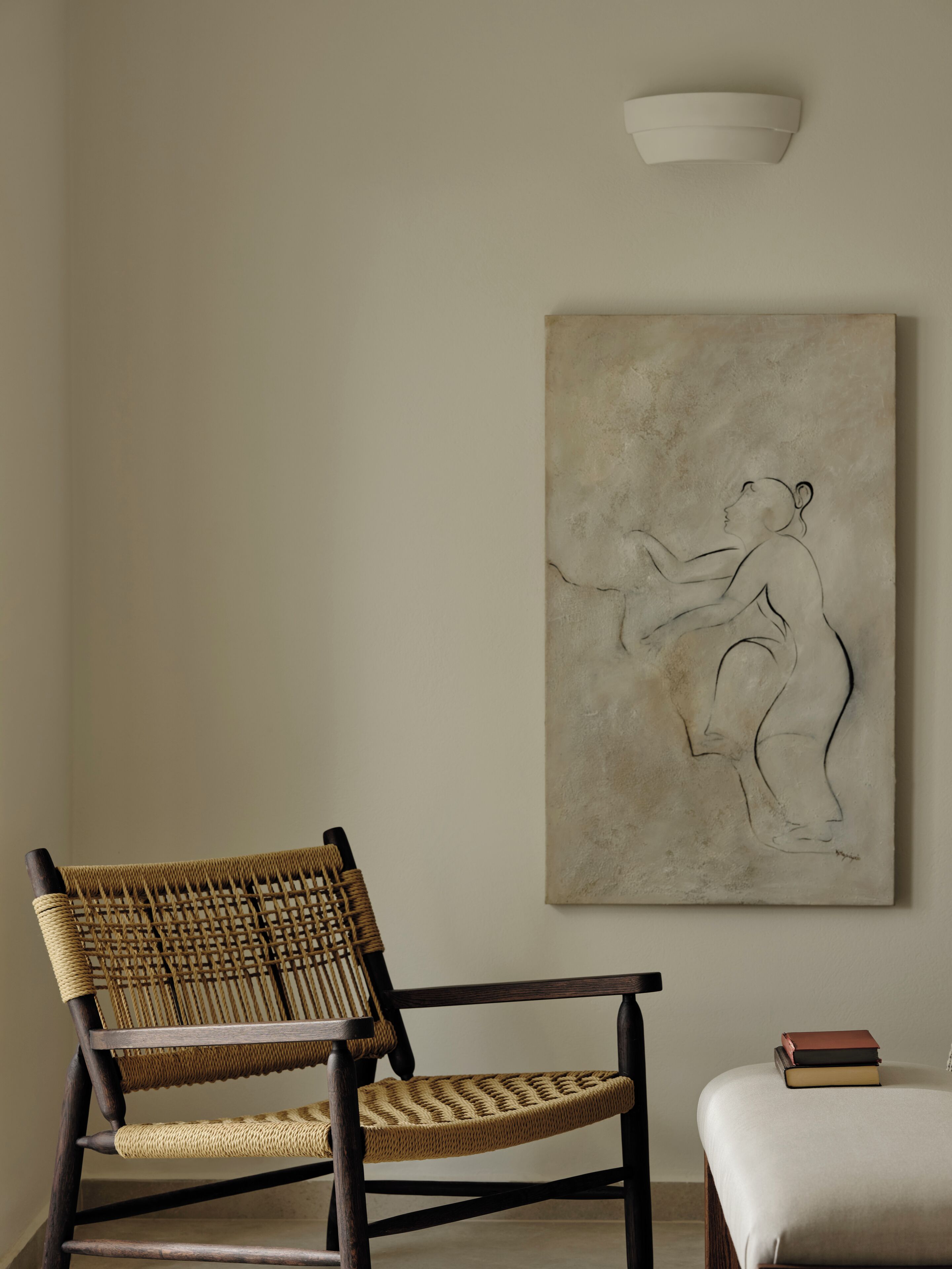 Una habitación con una silla de mimbre, un banco blanco acolchado y una obra de arte en la pared que representa una figura humana.