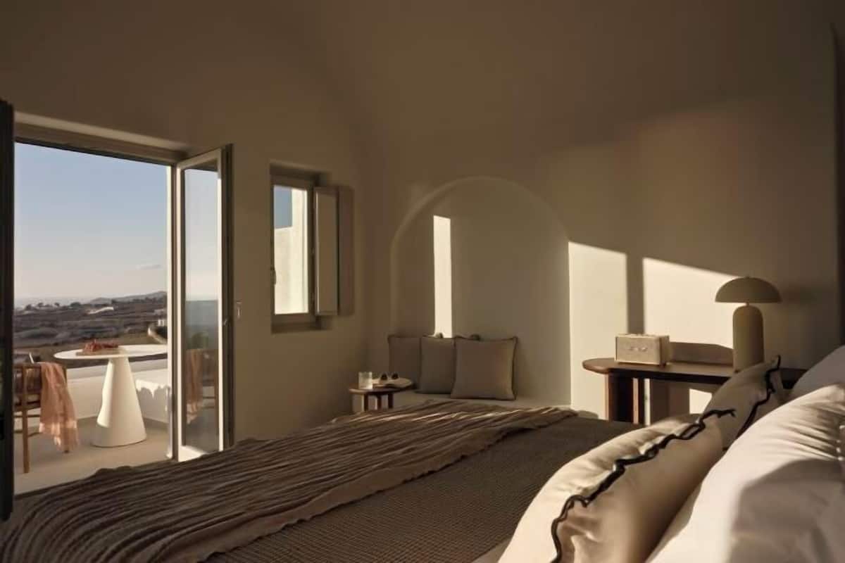 Quarto com cama, mesa de cabeceira, abajur, cadeira, mesa e vista através de uma janela grande.
