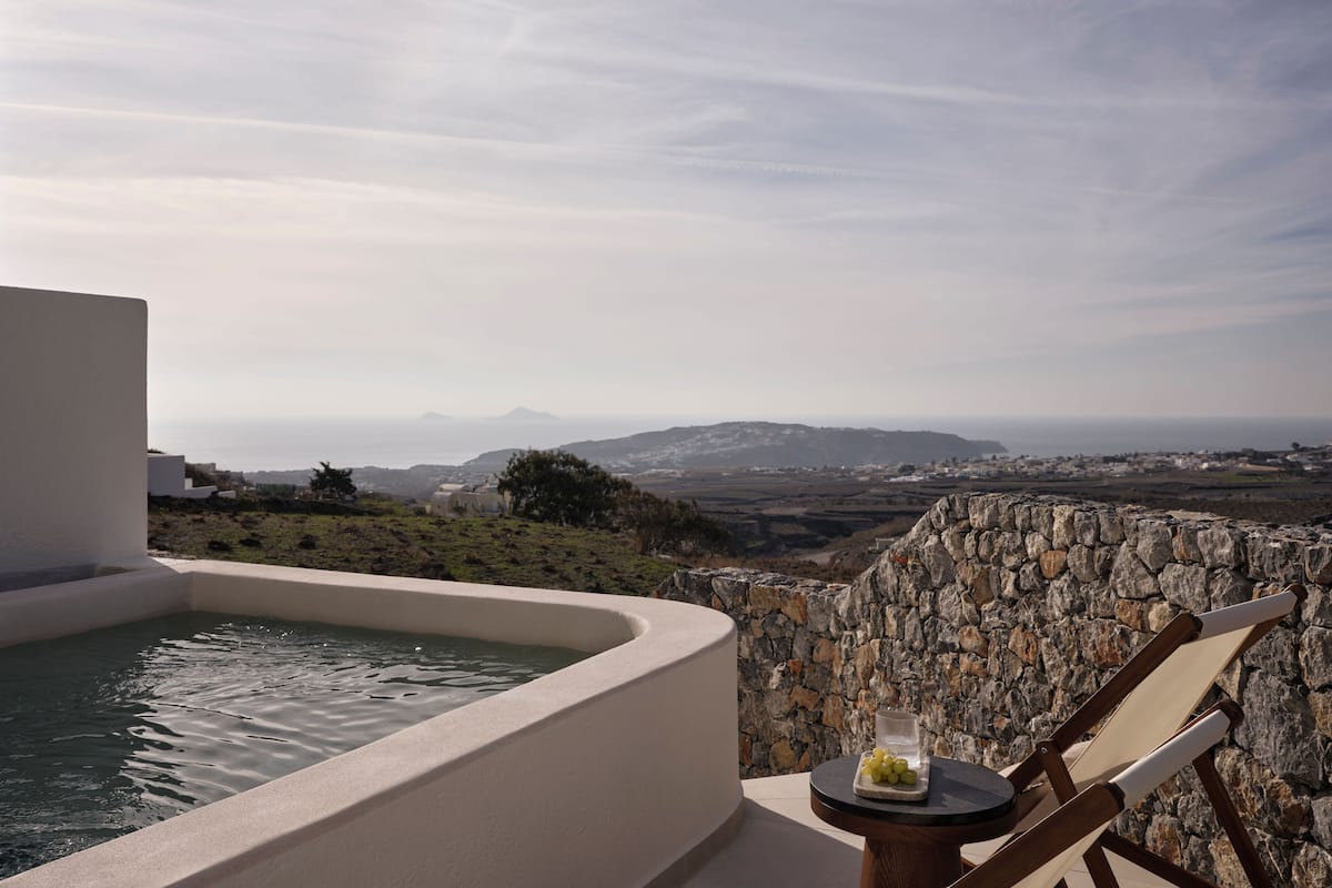 Uma banheira de hidromassagem com parede de pedra e mesa com copo e uvas, com vista para uma paisagem deslumbrante com montanhas e uma cidade.