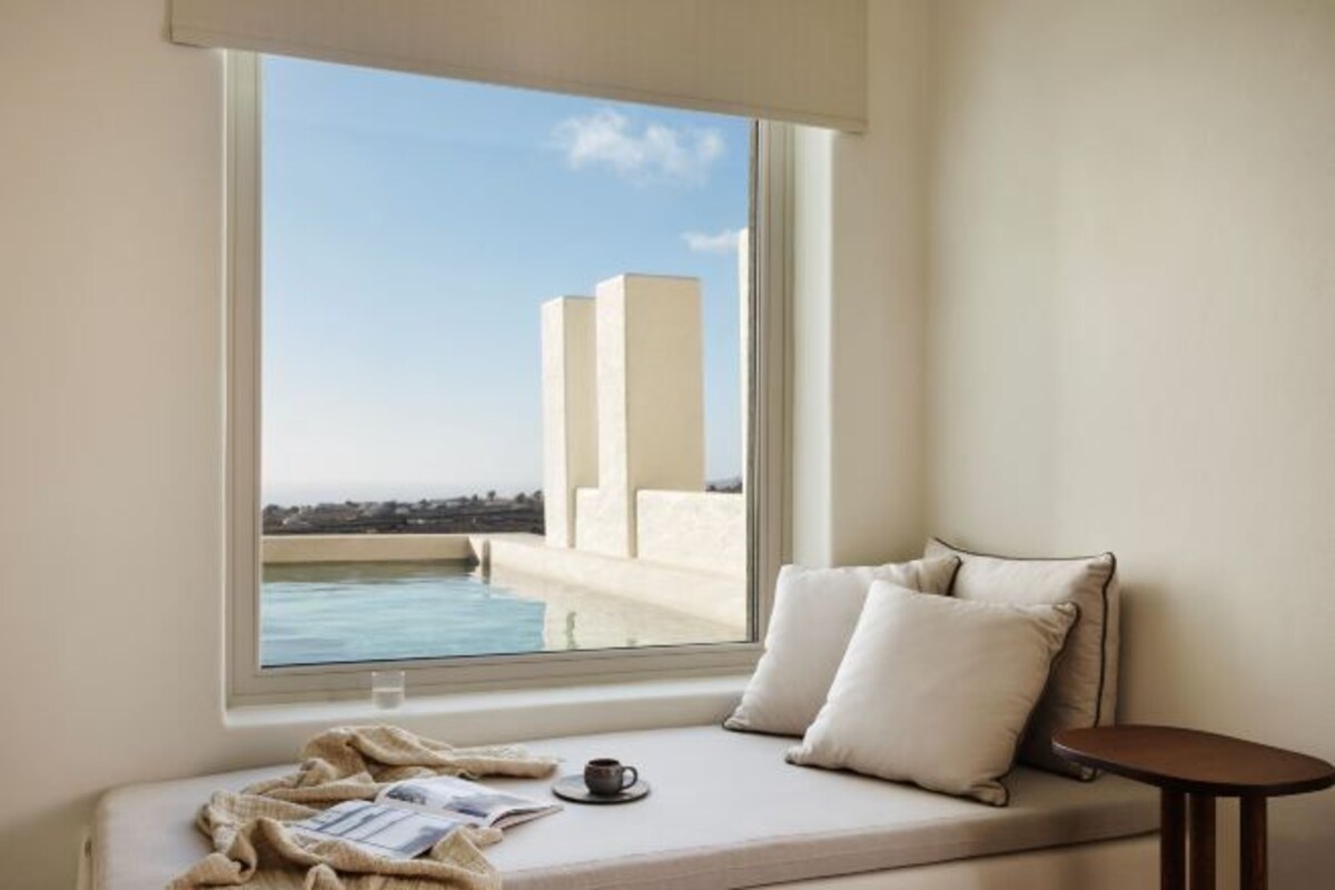 A room with a window overlooking a pool and buildings, a sofa with cushions, and a small table.