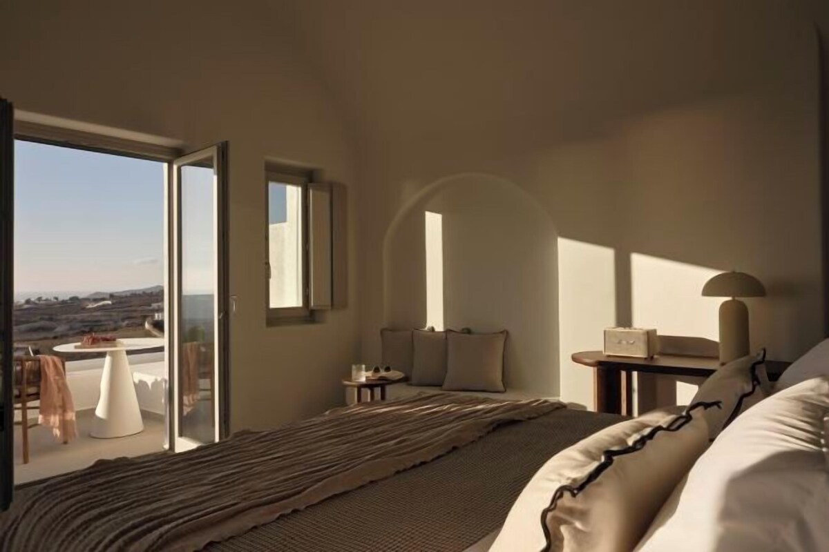 A bedroom with a bed, a nightstand, a lamp, a chair, a table, and a view through a large window.