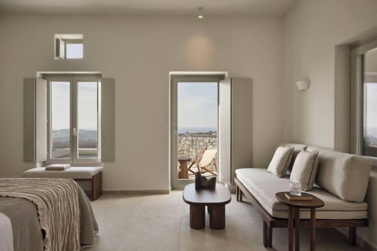 Quarto moderno com cama, sofá, mesinha e um banco. Duas janelas oferecem vista para o exterior.