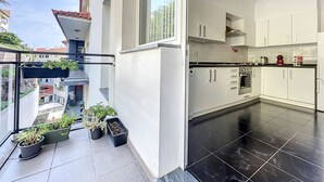 Una cucina moderna con mobili bianchi e pavimento nero, un balcone con piante in vaso e vista sugli edifici circostanti.