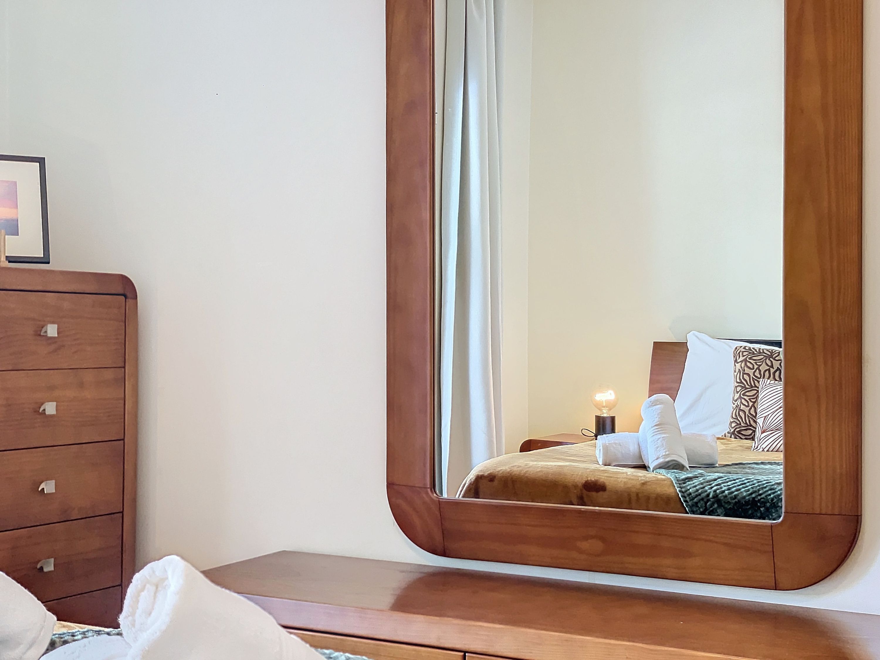 A bedroom with a wooden dresser, a large mirror, and a bedside table.