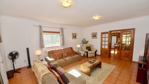 A living room with a sofa, armchair, and coffee table. There is a window with curtains, a fan, and a lamp. - Casa Bianca (A dos Francos)