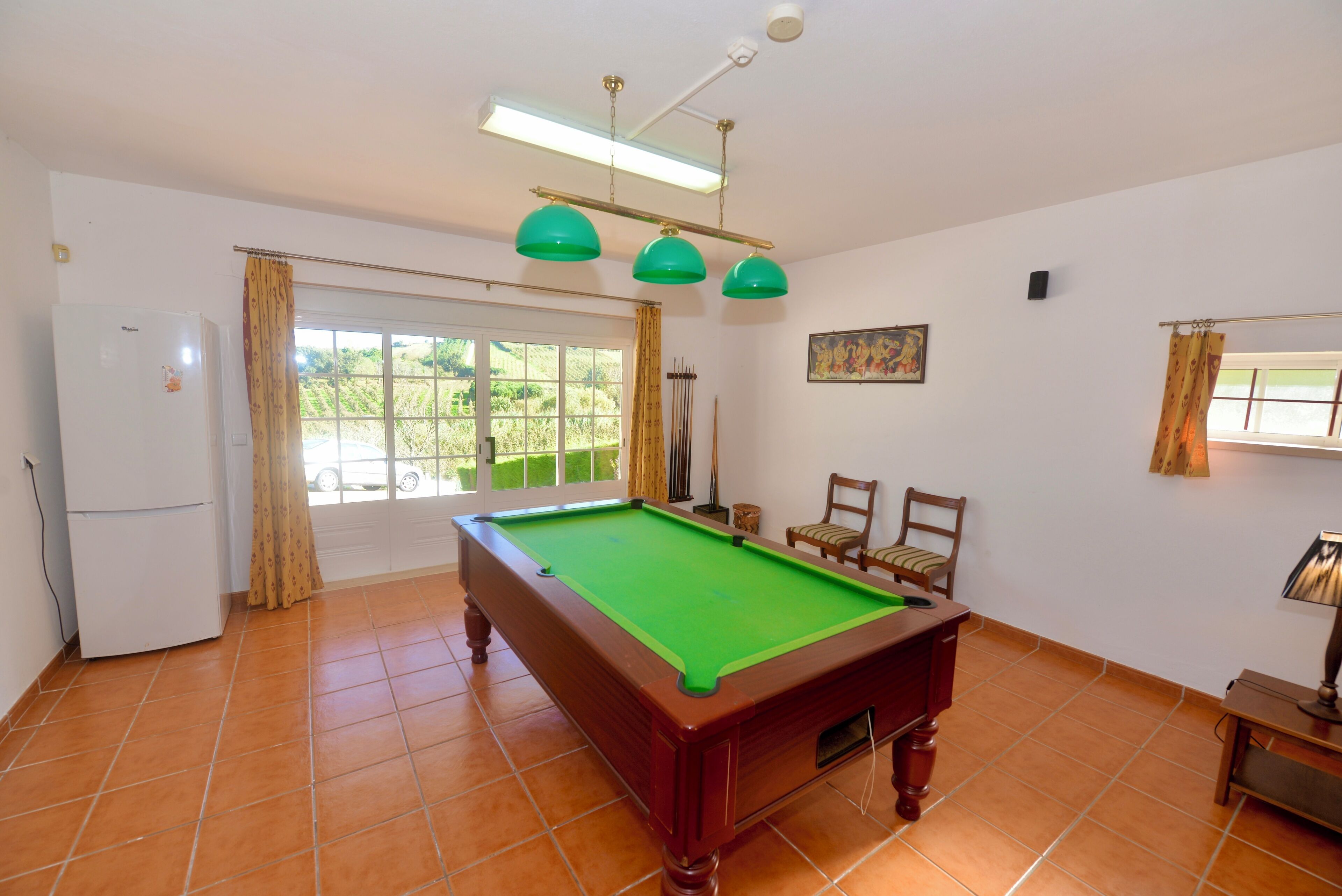 A pool table in a room with a refrigerator, chairs, a window with curtains, and a painting on the wall.