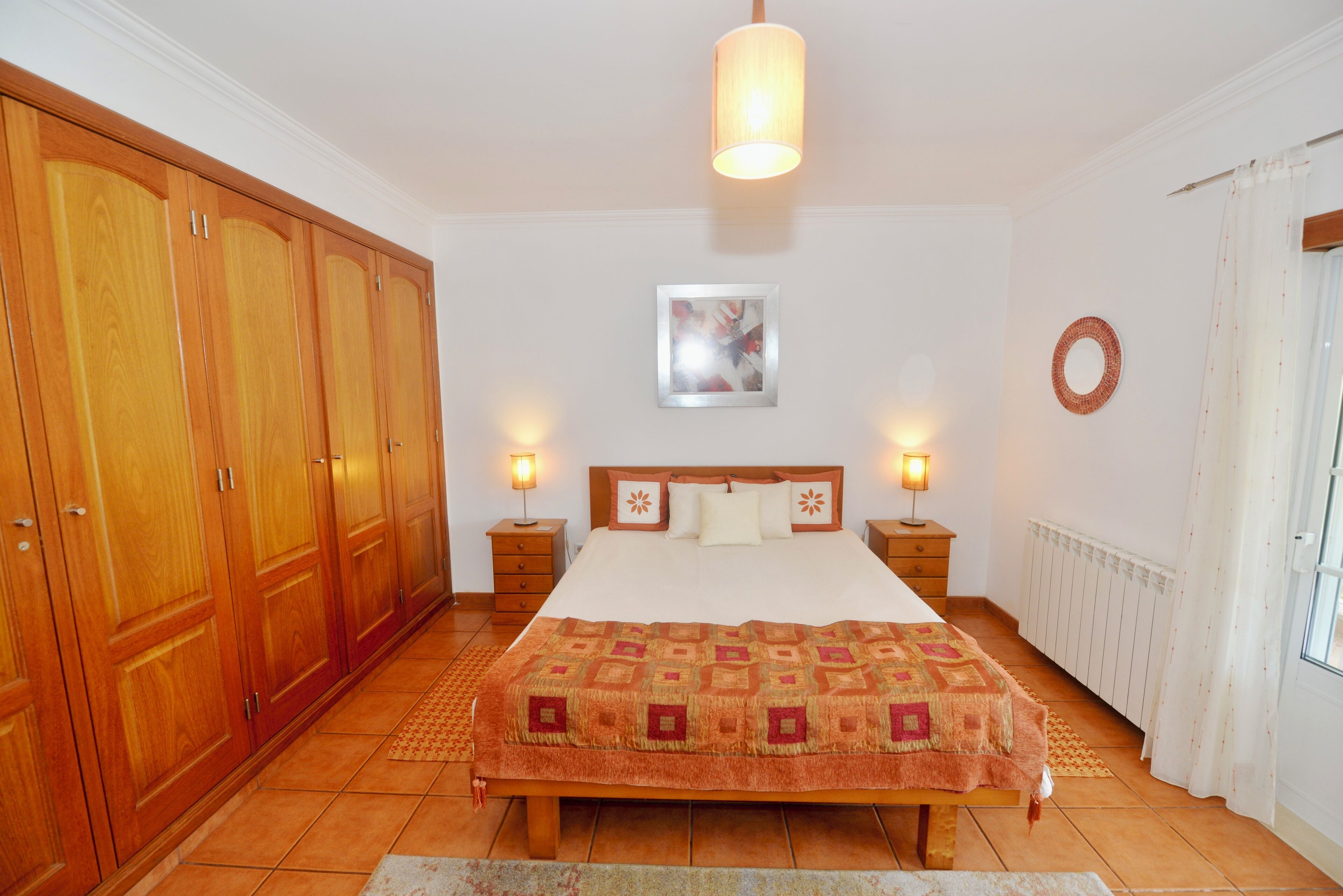 Un dormitorio con una cama grande, un armario de madera, dos mesitas de noche con lámparas, un cuadro enmarcado en la pared y un radiador.