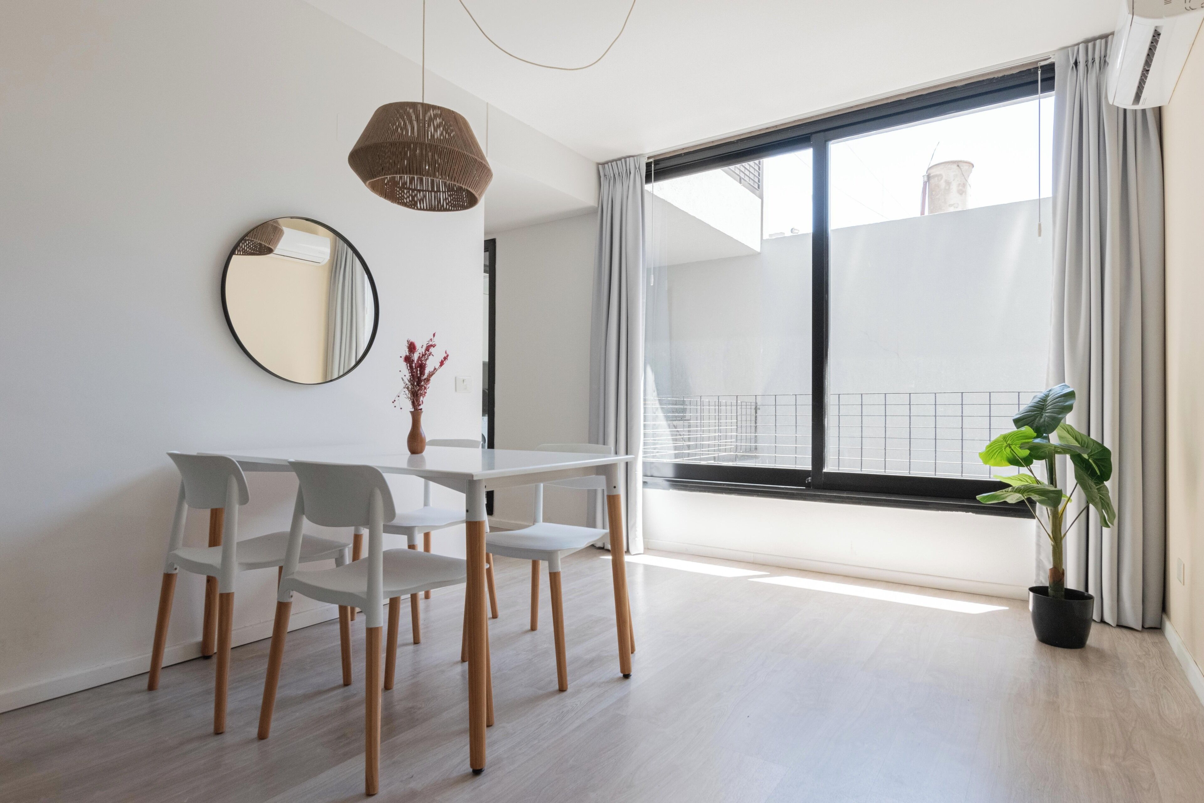 Ein moderner Essbereich mit einem weißen Tisch und Stühlen, einem runden Spiegel, einer Pendelleuchte und einem großen Fenster mit Blick auf den Himmel.