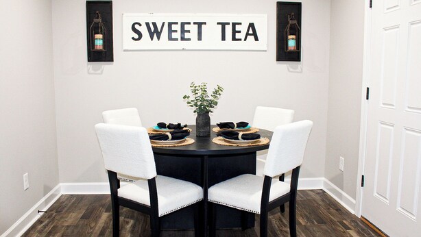 Un'area pranzo con un tavolo rotondo, quattro sedie bianche, un vaso con una pianta e un cartello appeso al muro con la scritta "SWEET TEA".
