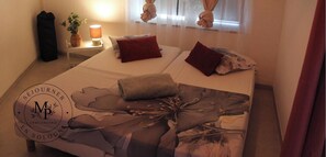 Hotelkamer met een bed, kussens, nachtkastje met een lamp, een plant en een raam met gordijnen.