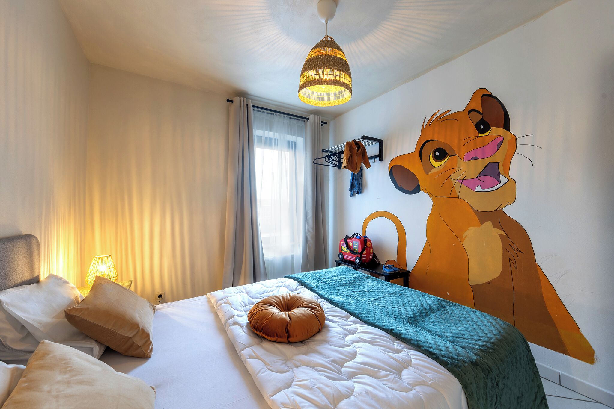 Een hotelkamer met een bed, een hanglamp, een wandkapstok en een grote muurschildering van een cartoonfiguur.