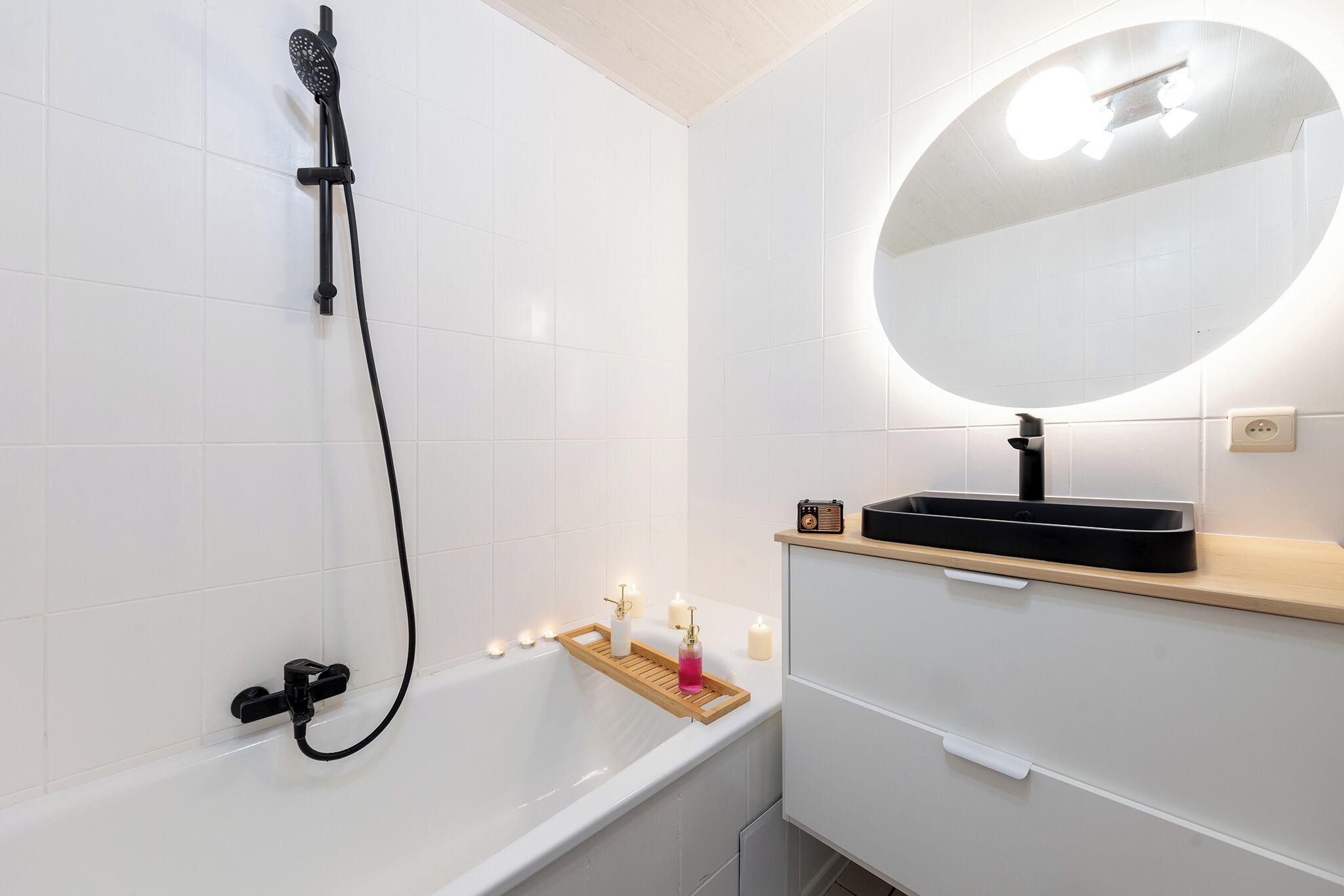 A modern bathroom with a white bathtub, a handheld showerhead, a vanity with a mirror, and a wooden tray holding candles.