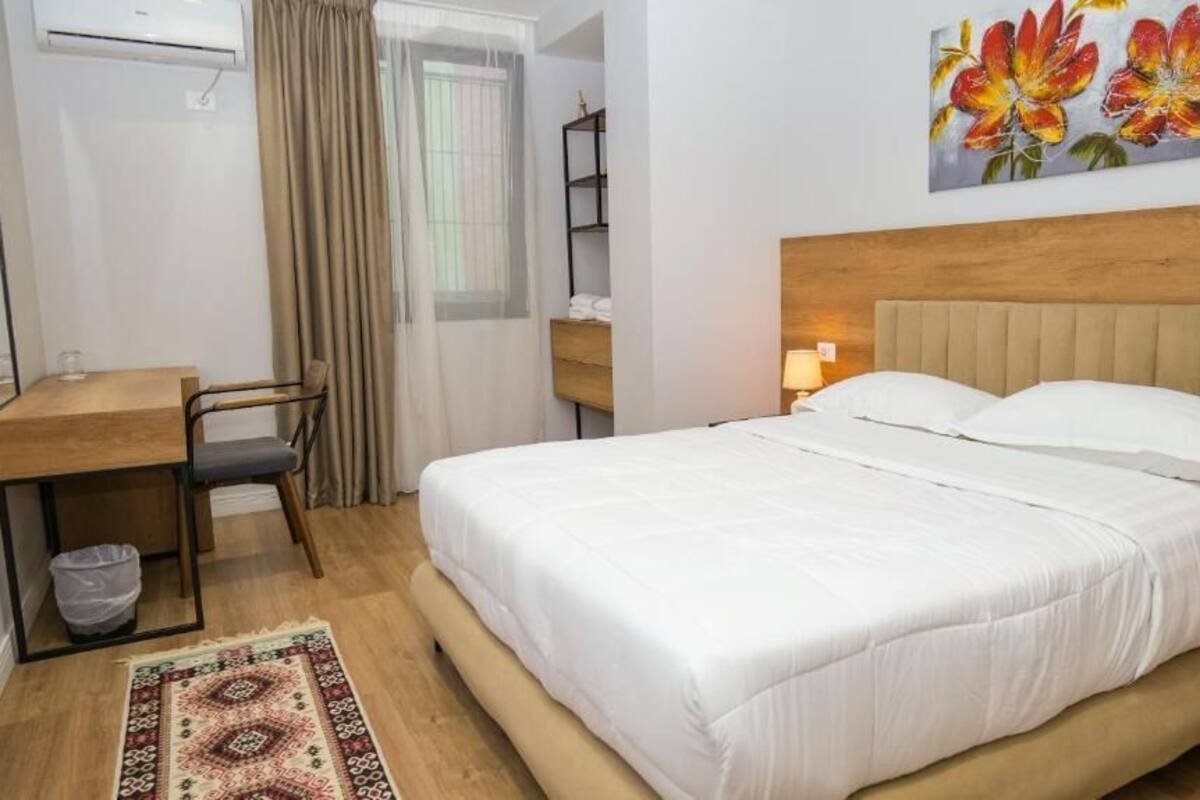 Une chambre d’hôtel avec un lit, un bureau, une chaise, une poubelle, une table de chevet et un tableau représentant des fleurs accroché au mur.