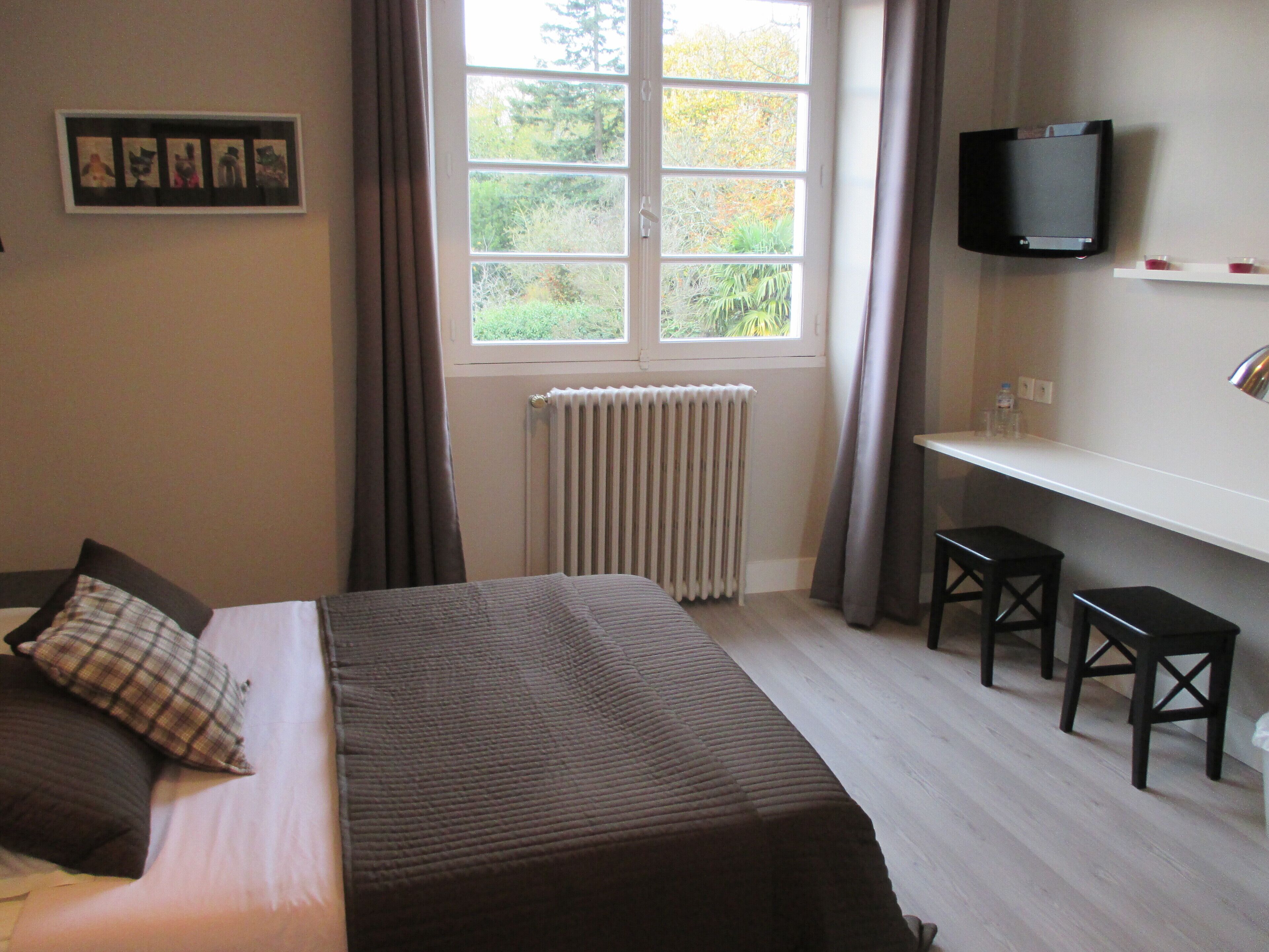 A bedroom with a bed, a desk with two chairs, a TV, and a window with curtains.