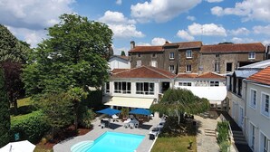 A swimming pool with lounge chairs and umbrellas, surrounded by a garden and buildings with red-tiled roofs.