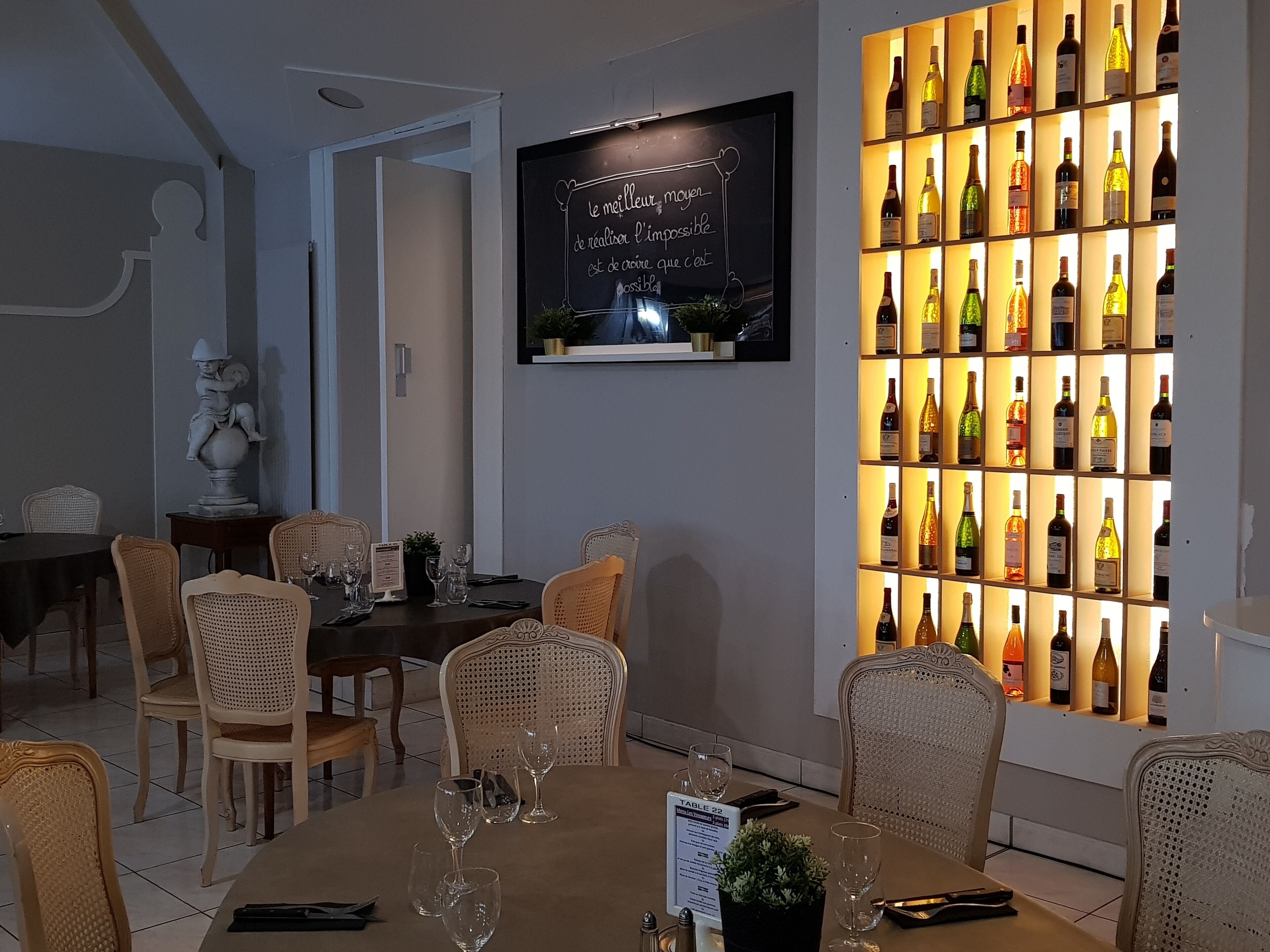 Un comedor con sillas de mimbre, una mesa dispuesta con copas de vino y una estantería de pared con botellas iluminadas.