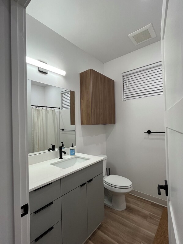 A modern bathroom with a white sink, mirror, and toilet. There is a wooden cabinet above the toilet and a window with blinds.