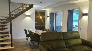 Un salon moderne avec un canapé vert, un plancher en bois, une table à manger avec des chaises, un escalier et une vue à travers une porte vitrée.