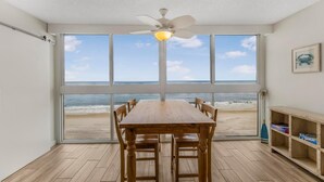 A dining area with a wooden table and chairs, large windows with a view of the beach, and a ceiling fan. - Edgewater West 65 (Gulf Shores)