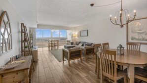 A spacious living area with a view of the beach, wooden furniture, and a chandelier. - Edgewater West 65 (Gulf Shores)
