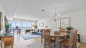 A spacious dining area with a wooden table and chairs, a chandelier, and a view of the ocean through large windows. - Edgewater West 65 (Gulf Shores)