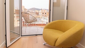 Una sedia gialla in una stanza con una porta vetrata che dà su un balcone con una poltrona in vimini e vista su edifici e colline.