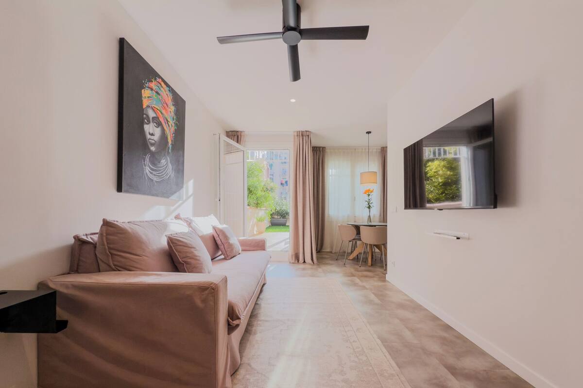 A modern living room with a sofa, a ceiling fan, a TV, and a view of the outdoors.