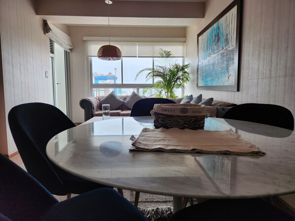 A modern dining area with a round glass table, dark chairs, a woven basket with a label, a large window with a city view, and a hanging light fixture.