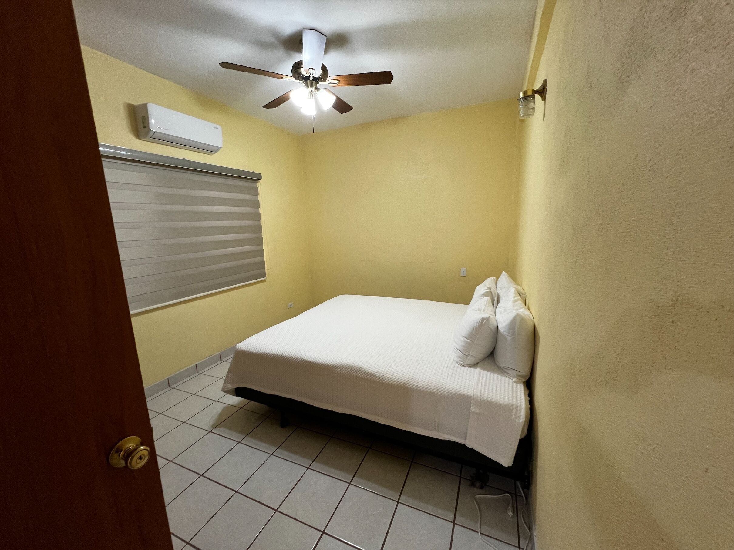 Una habitación pequeña y limpia, con cama, ventilador de techo y aire acondicionado.