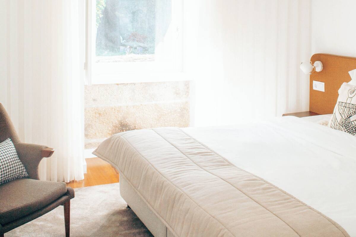 Une chambre à coucher comprenant un lit, une chaise, un luminaire suspendu et une fenêtre avec des rideaux.