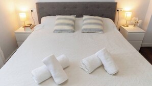 Ein ordentlich bezogenes Bett mit zwei Kissen, zwei aufgerollten Handtüchern und je einer Nachttischlampe auf weißen Nachttischen.