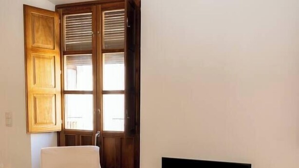 A room with a wooden floor, a white chair, a television on a white stand, and a window with wooden shutters. - Puerta Real Apartments (Granada)