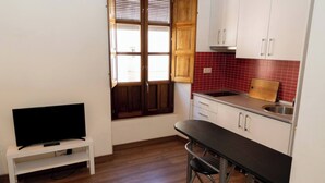 A compact kitchen with a wooden window, white cabinets, a red-tiled backsplash, a black countertop, and a small TV on a white shelf. - Puerta Real Apartments (Granada)