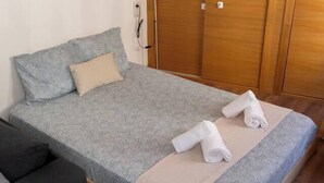 A single bed with a grey patterned bedspread, a beige pillow, and two rolled towels on the bed. There is a wooden wardrobe with a mirror and a lamp on the wall. - Puerta Real Apartments (Granada)
