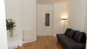 A room with a wooden floor, a grey sofa, a white radiator, and a wall-mounted lamp. - To complete (Granada)