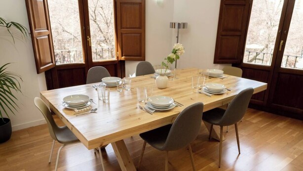 A dining room with a wooden table set for six, surrounded by six chairs, with a potted plant and a vase of flowers on the table. - To complete (Granada)