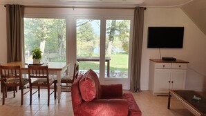 Een woonkamer met een rode bank, een eettafel met stoelen, een televisie aan de muur en uitzicht op een tuin door grote glazen deuren.