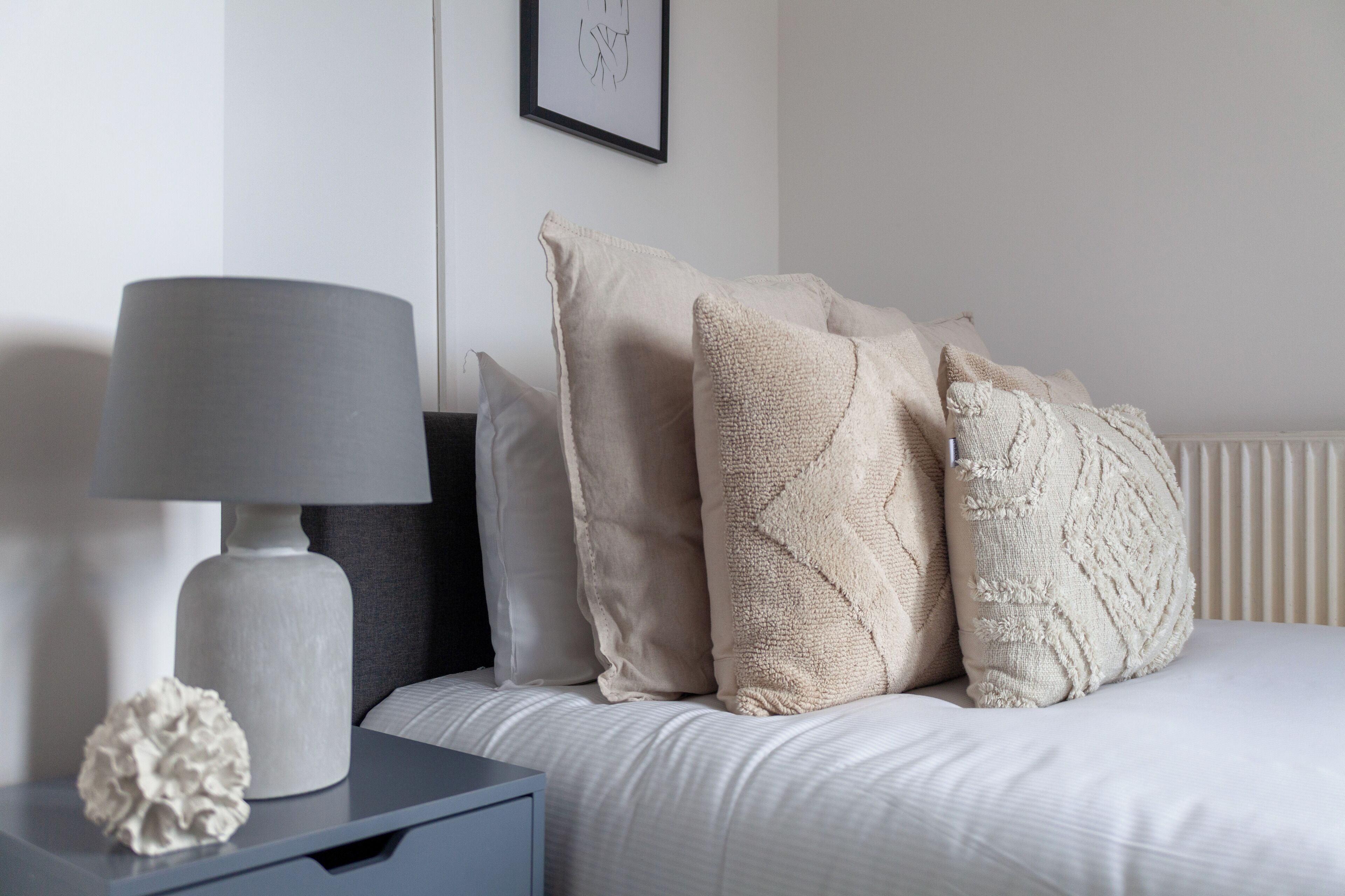 Un letto rifatto con un comofort bianco e diversi cuscini beige. Una lampada grigia è posizionata su un comodino accanto al letto.