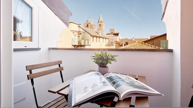 Un balcón con un banco de madera, una planta en maceta y una revista abierta. Se observa un edificio con una torre al fondo.