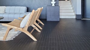 Ein modernes Wohnzimmer mit einem grauen Sofa, Korbstühlen, einem kleinen Beistelltisch mit Lampe und einer Treppe, die in ein Obergeschoss führt.