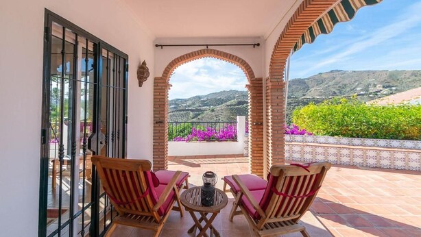 Un balcón con dos sillas de madera y una mesa, con vistas a un jardín de flores púrpuras y un paisaje montañoso.