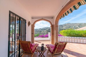 Un balcón con dos sillas de madera y una mesa, con vistas a un jardín de flores púrpuras y un paisaje montañoso.