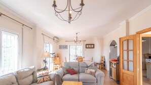 Amplia sala de estar con un sofá gris, una mesa de centro de madera y una araña. Varias ventanas ofrecen luz natural y vistas de la ciudad.