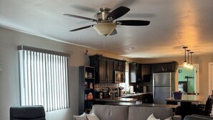 Una sala de estar moderna con un sofá gris, dos sillones de cuero y un ventilador de techo. Se ve una cocina con armarios oscuros y un refrigerador al fondo.