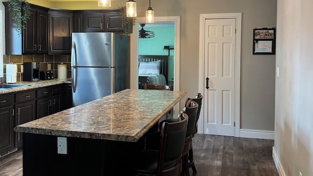 Una cocina con armarios de madera oscura, un refrigerador de acero inoxidable, una isla de granito con taburetes de bar, iluminación colgante y vistas al comedor contiguo.