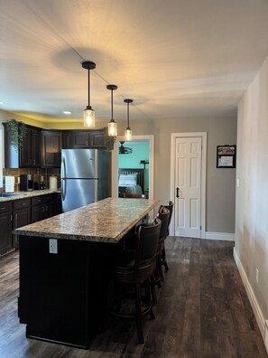 A kitchen with dark wood cabinets, a stainless steel refrigerator, a granite countertop island with bar stools, pendant lighting, and a view into the adjacent dining area.
