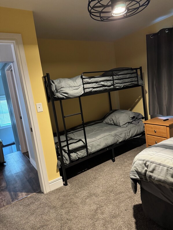 A bunk bed with a wooden nightstand and a small table, a door leading to another room, and a ceiling light fixture.