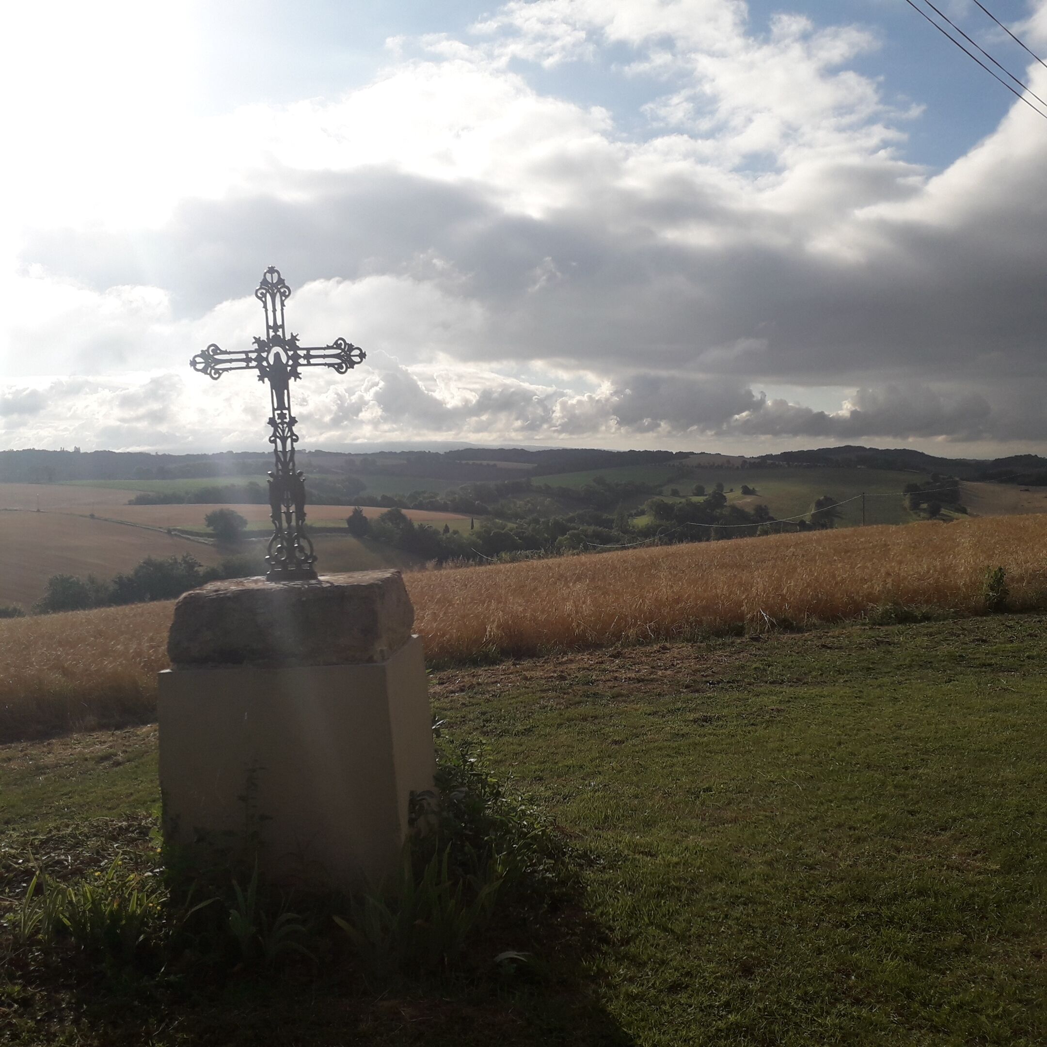 A rural landscape with a cross monument in the foreground and a field in the background.